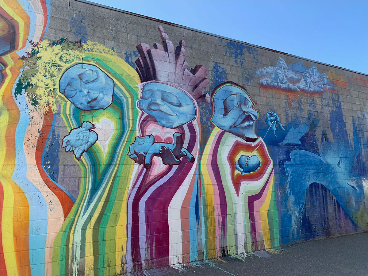 A large mural spans a cement-block wall below a blue sky. Three illustrated figures emanate light and offer a dove, a baby and a heart stitched together with thread.