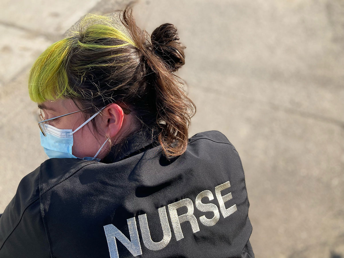 A person facing away from the camera wears a black jacket that reads ‘NURSE’ in bold white letters across the back. Their face is turned to the side, showing green bangs, a medical mask and glasses. There is faded pavement in the background.