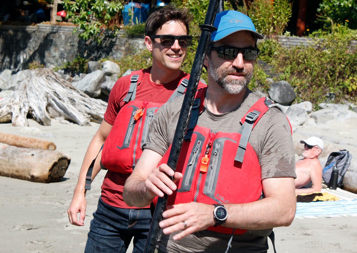 Two men stand in lifejackets and sunglasses on a West Vancouver beach. One holds a kayak paddle.