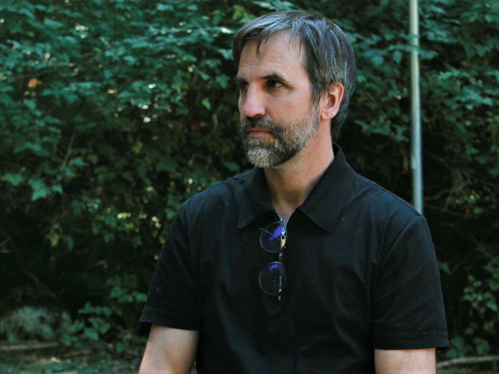 A dark haired, bearded white man with flecks of gray, federal environment Minister Steven Guilbeault, wears a dark short sleeved shirt with sun glasses hanging from his neck, looking to his right in front of foliage.