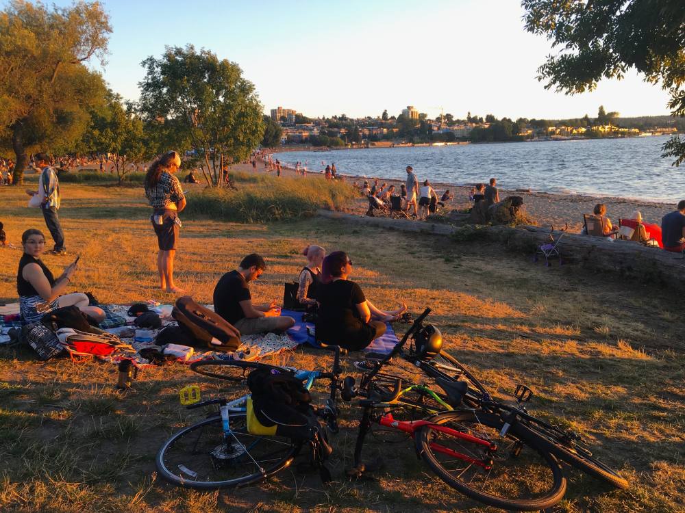 People are enjoying the sunset on picnic blankets at Vancouver’s Kitsilano Beach. They’ve ridden their bikes here, which are laying down in the sand.