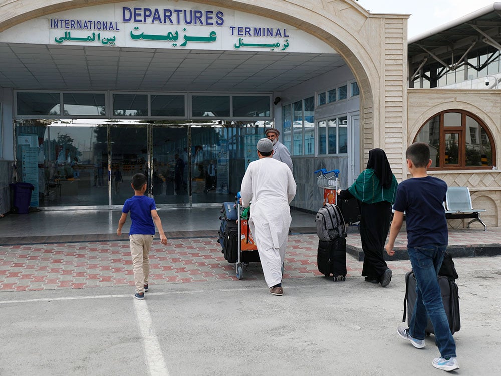 A family approaches the glass doors of a building. Above the doors, a sign reads in English and Arabic, “International Departures Terminal.”
