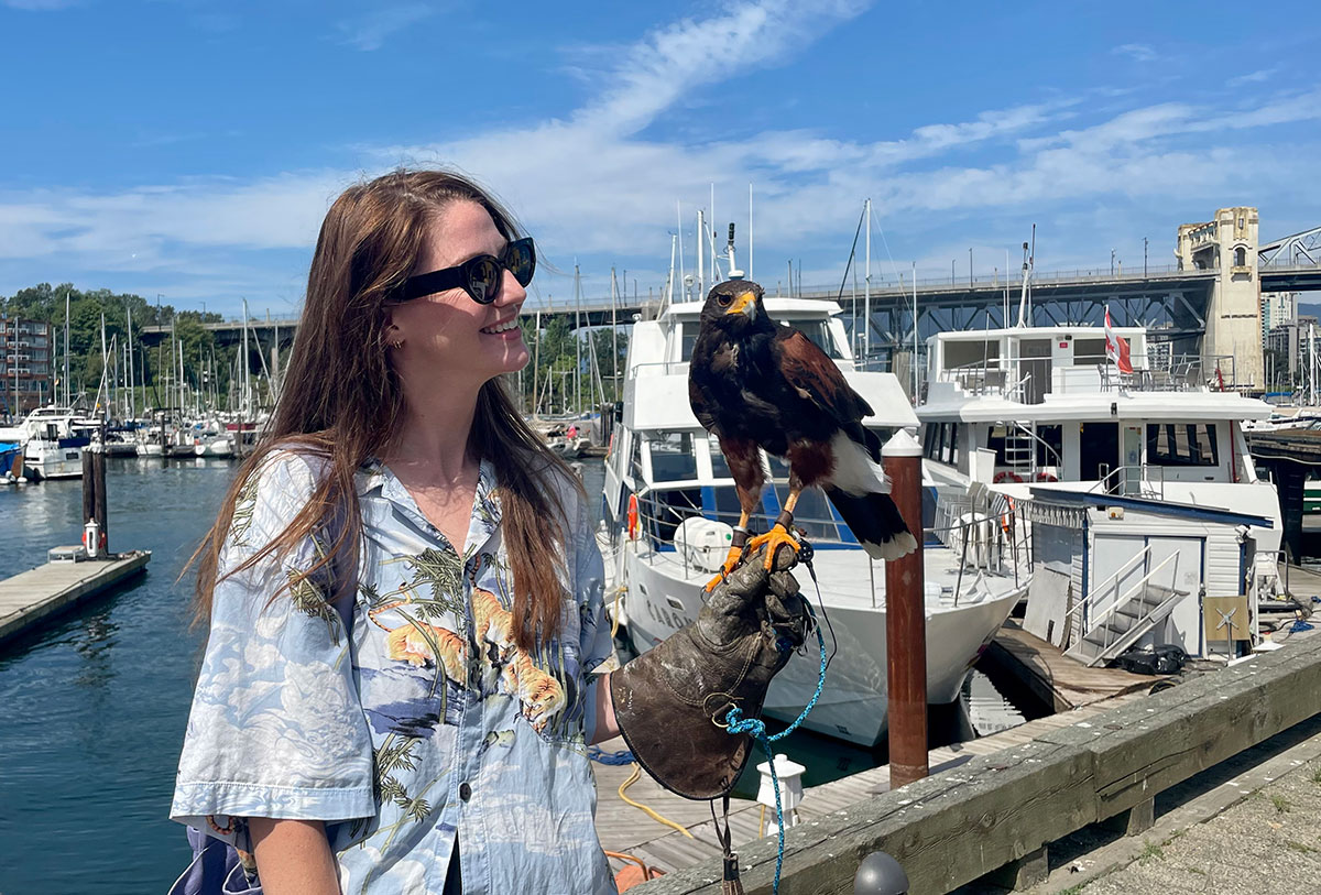 The author stands on Granville Island. She is wearing a leather glove. A Harris’s hawk sits on the glove.