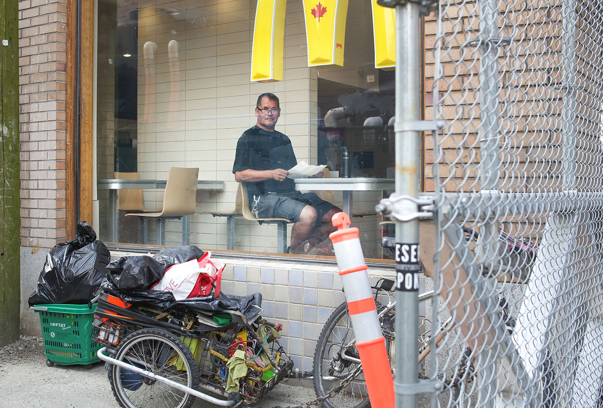 The view inside of a McDonald’s from the street. There is a man sitting by the table in the window, looking at the camera, holding a document. He has a bike and trailer parked outside the window, lugging several plastic bags.