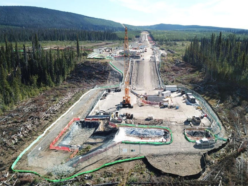 An aerial view shows a large cleared, graded area carved out of the forest. Equipment, including a towering yellow drilling rig, is in the foreground as the pipeline route stretches into the distance.