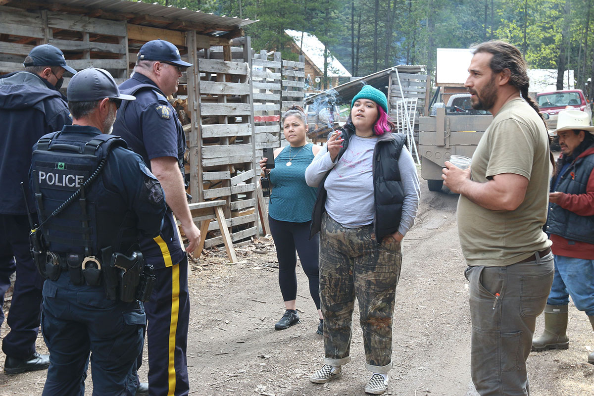 Three RCMP uniformed RCMP officers face two women and two men on a muddy road. A shelter made of old pallets is on the left. Two trucks are in the background.