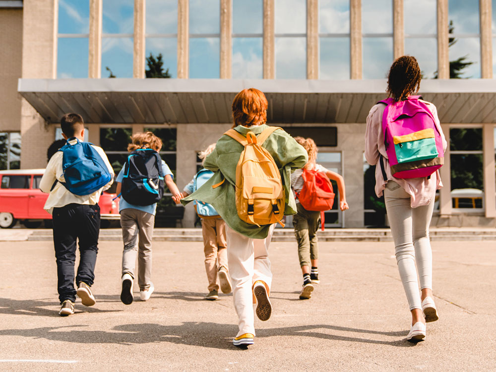 Kids enthusiastically make their way towards a school entrance, wearing backpacks.