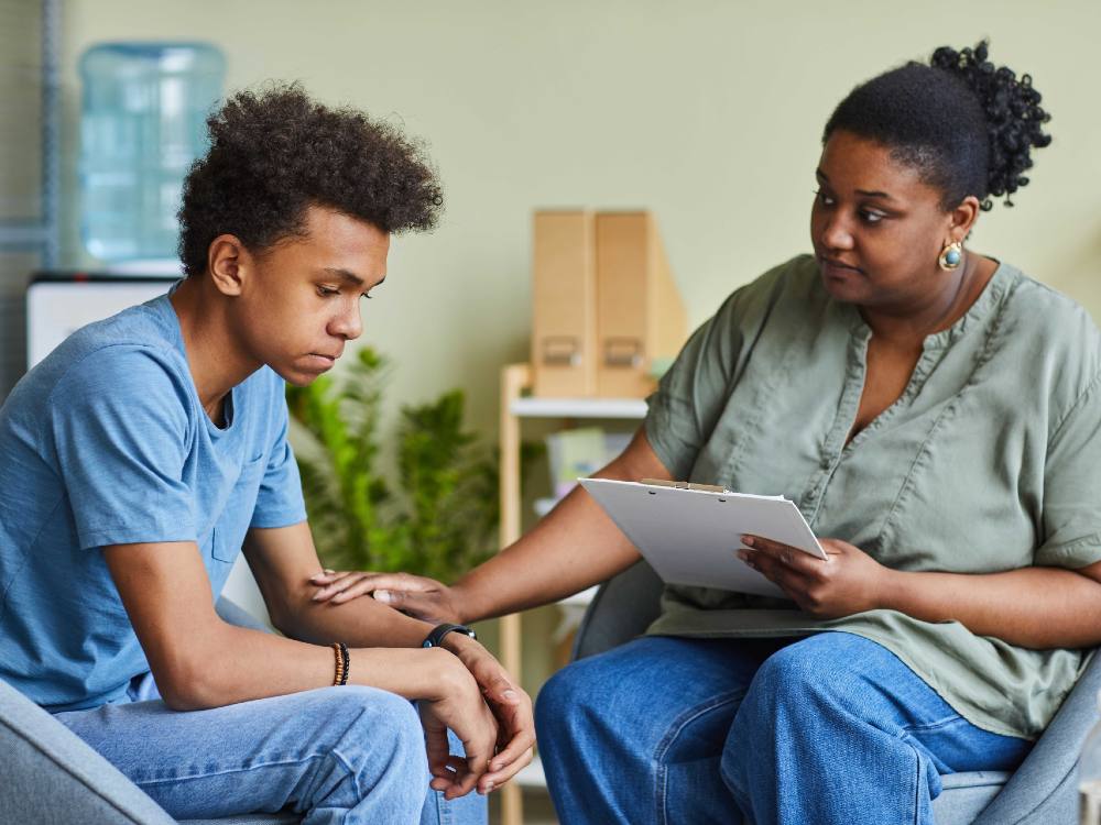 A Black teenage boy is seated to the left of a frame in a blue T-shirt and jeans. A Black woman to his right places her hand on his arm in a supportive gesture. She is wearing medical scrubs. 