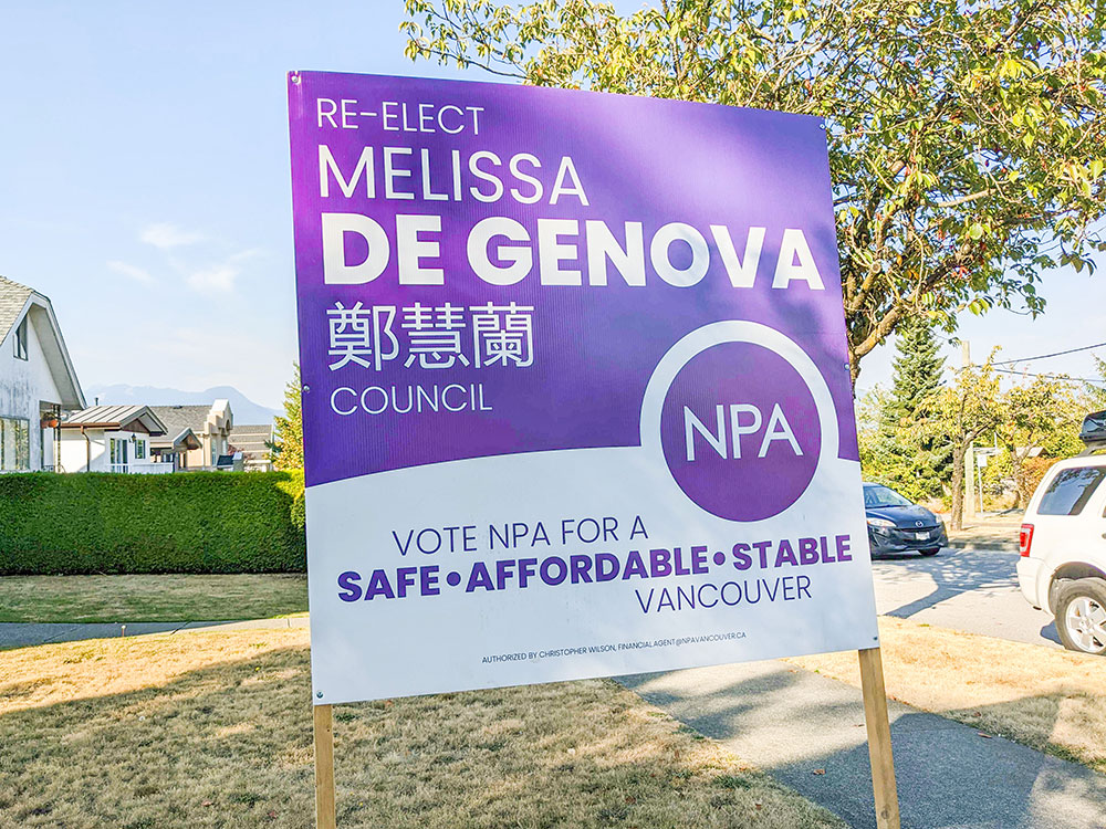 A lawn sign on a sunny day. It’s promoting the NPA’s Melissa De Genova for council, with the party’s signature purple colours. It features De Genova’s name in traditional Chinese characters.