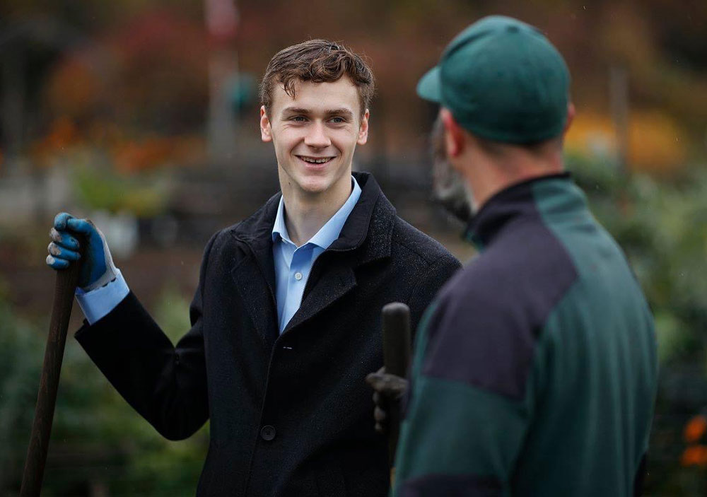 Ned Taylor is wearing a black peacoat over a light blue button-up shirt. He is wearing work gloves and holding the handle of a shovel or rake. He is talking to a person in the foreground whose back is turned to the camera. They are wearing a green ball cap and navy jacket.