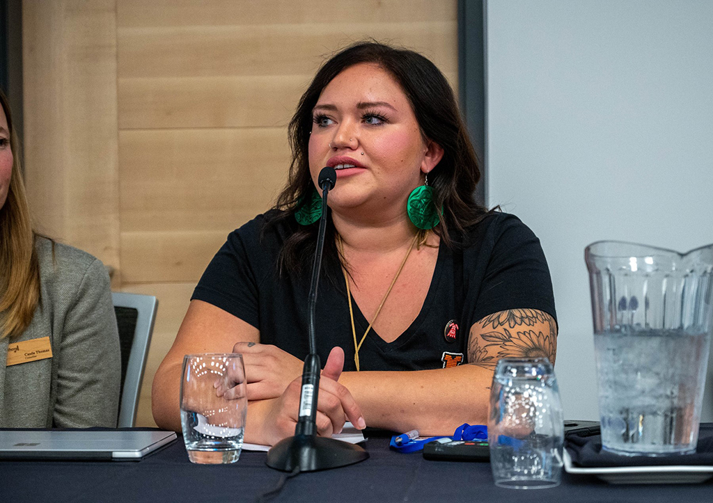 An Indigenous woman sits at a table, with dark hair and a dark short-sleeved short and large green earrings. She is speaking into a microphone.