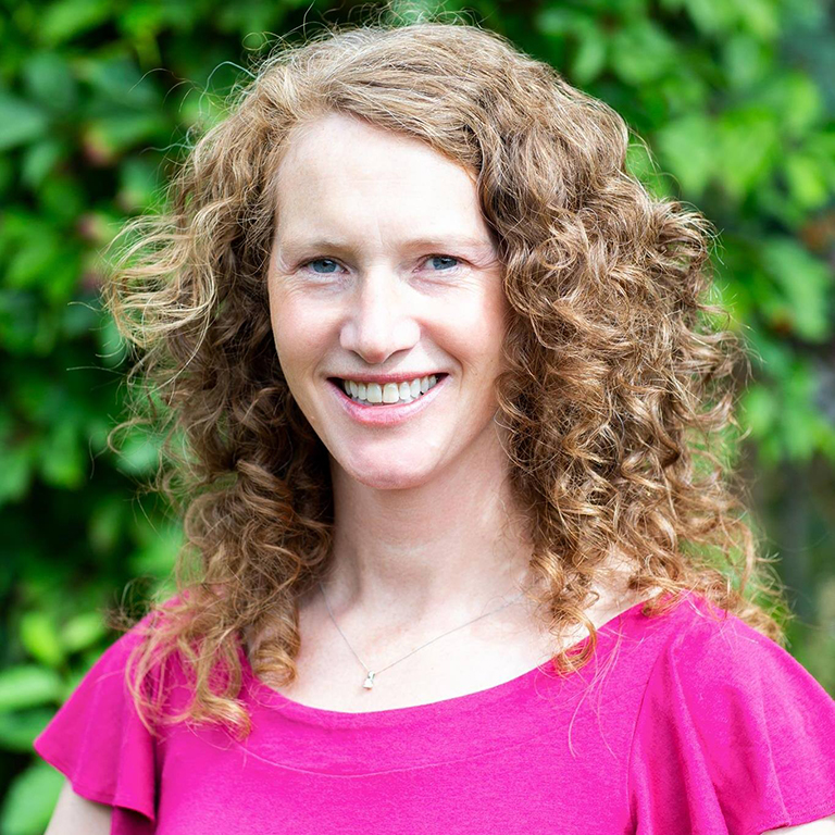 A woman with shoulder-length curly hair and a dark pink shirt smiles at the camera.