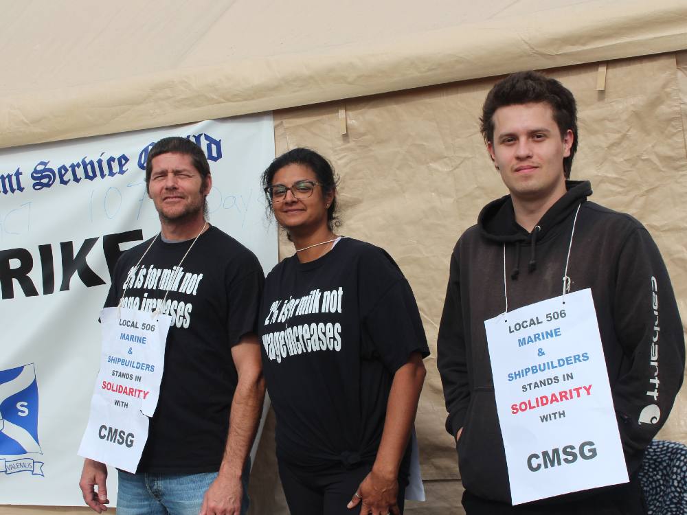 Three workers in black shirts — two white men and a woman who is brown, smile at the camera. Their shirts say “Two per cent is for milk, not wage increases,” and their signs identify them as members of Local 506 Marine and Shipbuilders who stand in solidarity with union CMSG.