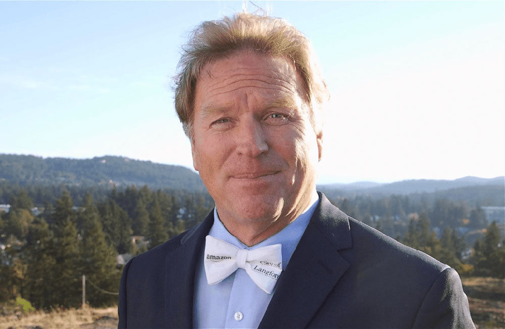 A brown-haired middle aged white man in a blazer and wearing a white bow tie with the words Amazon and Langford on it.
