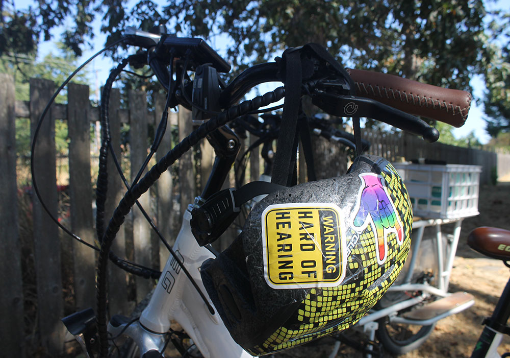 Naomi Wilde’s bike helmet hangs on the handlebars of her white bike. The helmet is adorned with stickers, including one in yellow and black text that reads 'Warning: hard of hearing.'
