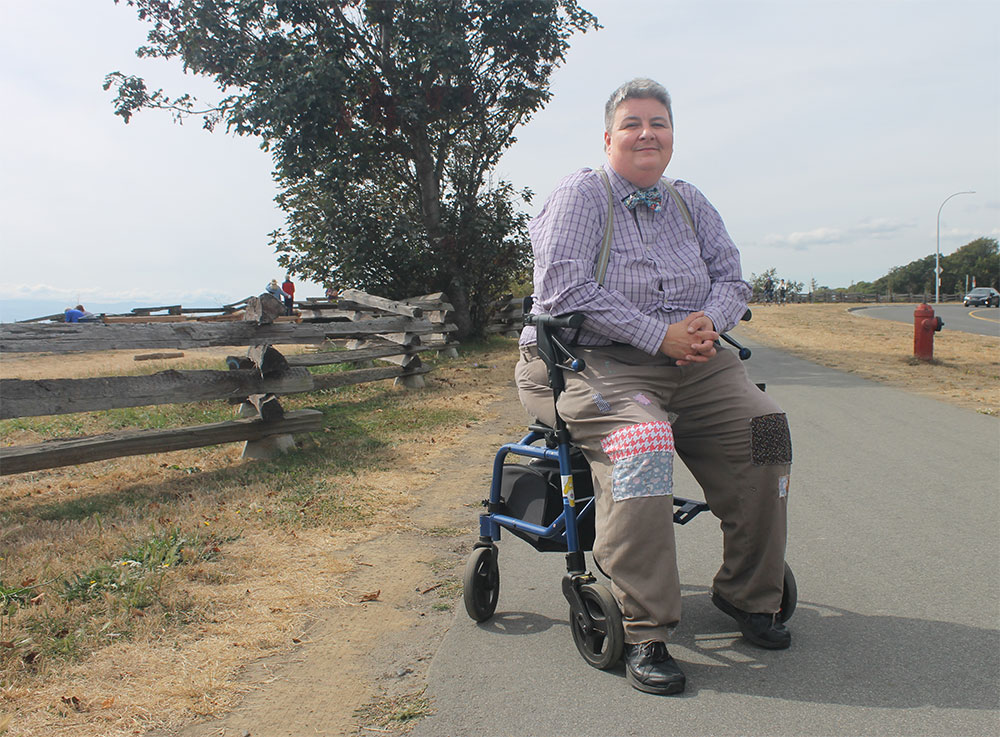 JJ Jones has short grey hair and is wearing a checkered purple button-down with a bow tie. They are sitting on their blue walker on a protected bike path near a beach.