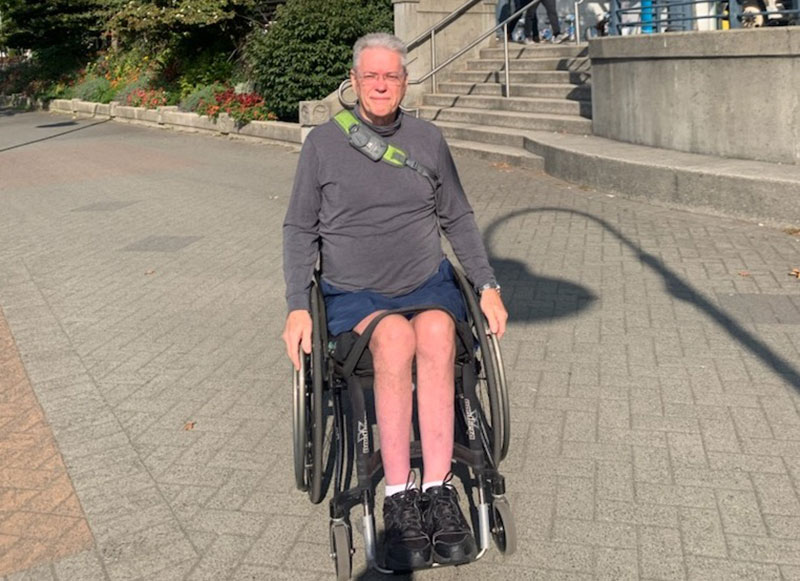 Jacques Courteau is wearing a grey shirt and shorts. He is in his wheelchair on Vancouver’s downtown seawall. He has grey hair and glasses and is looking directly at the camera.