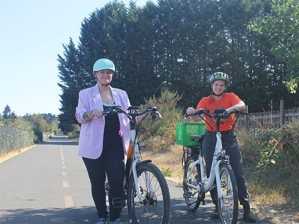 Two people stand with their bikes on a protected bike lane in Victoria. Kris Westendorp, left, is wearing a lilac blazer and light turquoise bike helmet. Naomi Wilde, right, is wearing an orange t-shirt and green bike helmet. They are both standing near with their white bikes and smiling at the camera. Stands of trees are in the background.
