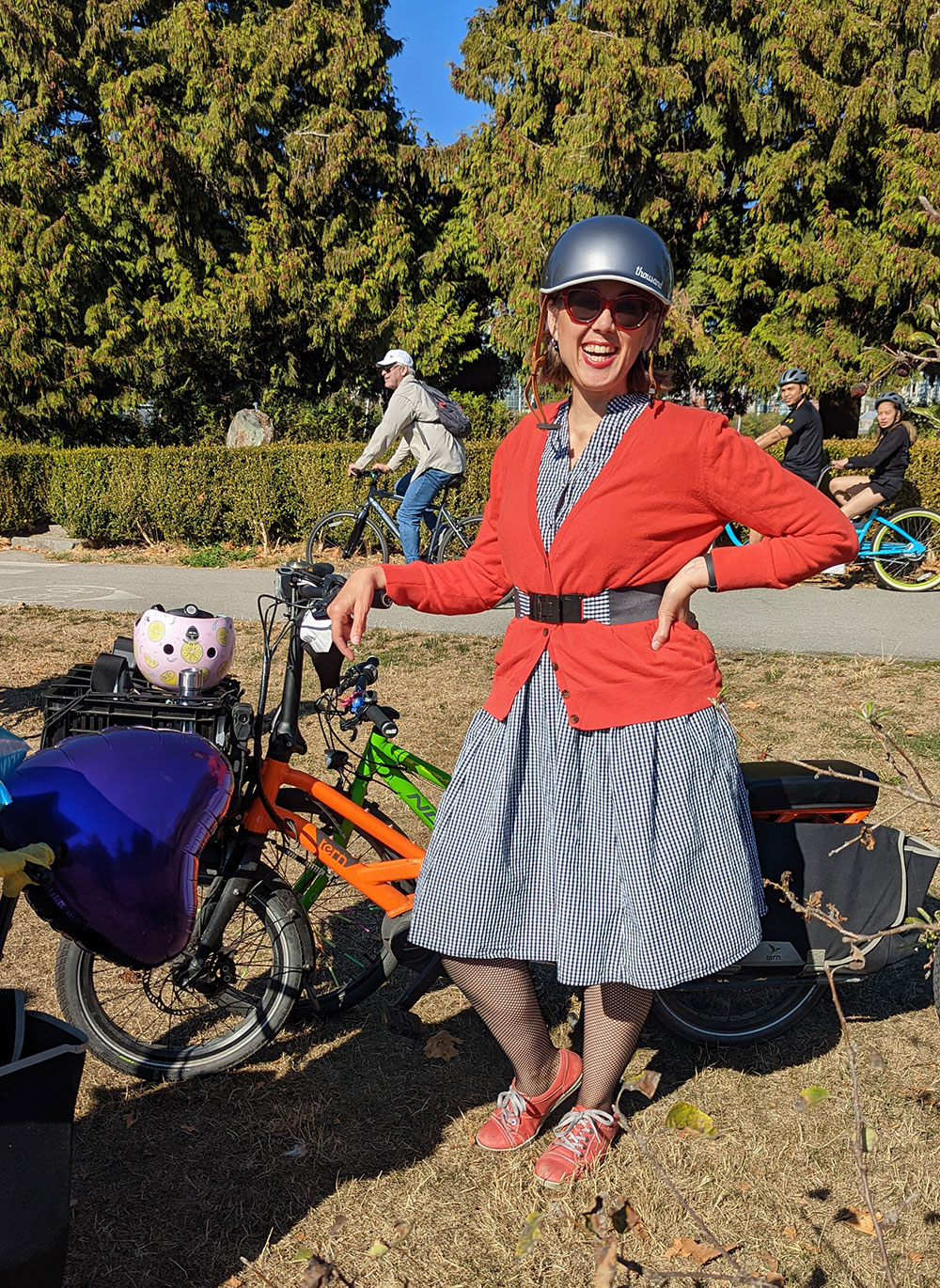 Lisa Corriveau is standing next to her bike with a hand on her hip, smiling. She is wearing sunglasses, a navy bike helmet, and an orange cardigan over a striped navy and white dress. Behind her are cyclists riding on a protected bike lane.