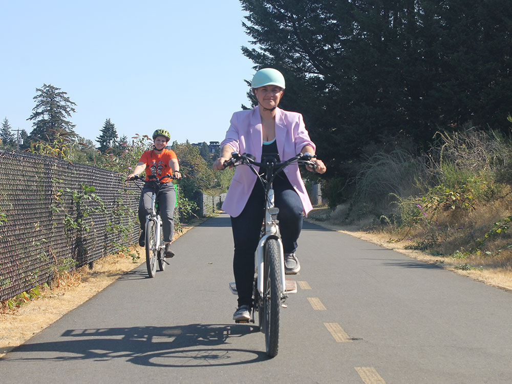 Kris Westendorp is riding her bike in the centre of the frame. She’s wearing a lilac blazer. Her coworker Naomi Wilde is wearing an orange t-shirt, riding behind Westendorp to the left of the frame. They are on a protected bike lane and smiling.