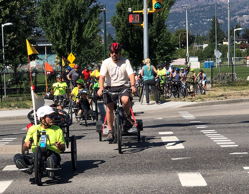 A crowd of handcyclists and cyclists cross an intersection. Many, including Paul Clark on the far left, are wearing neon green t-shirts. They are participating in a memorial event for a handcyclist killed at this intersection.