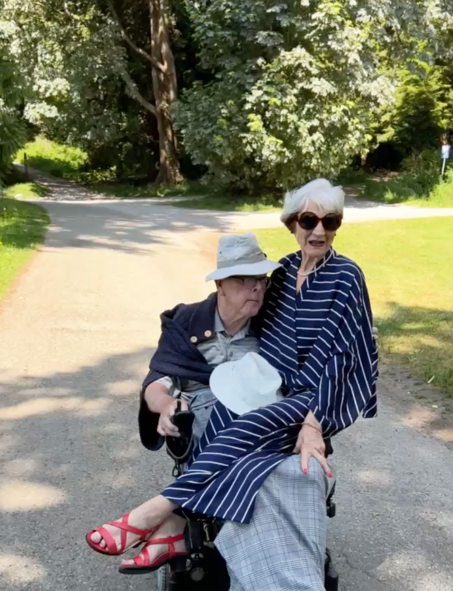 Peter and Janice Foran ride down a protected bike lane. Janice is wearing a navy dress and sunglasses, sitting on Peter’s lap. Behind them are trees and green grass.