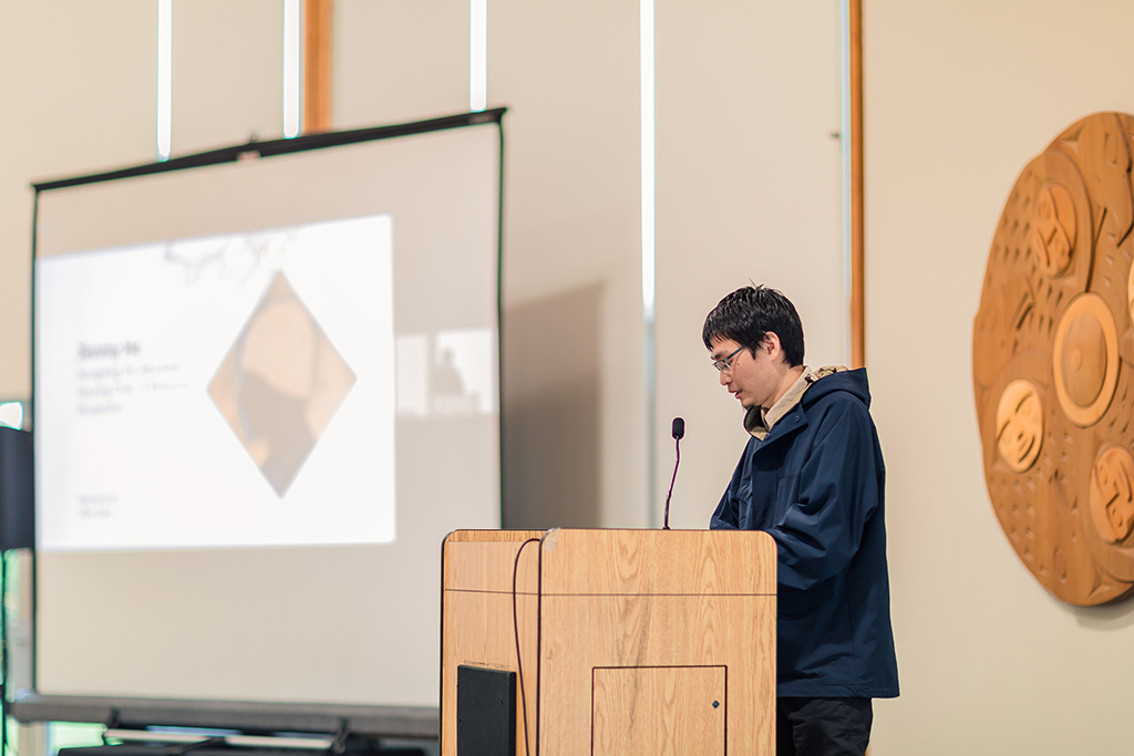 A young man wearing glasses stands at the podium. In the background, a screen with a slide from his presentation is visible.