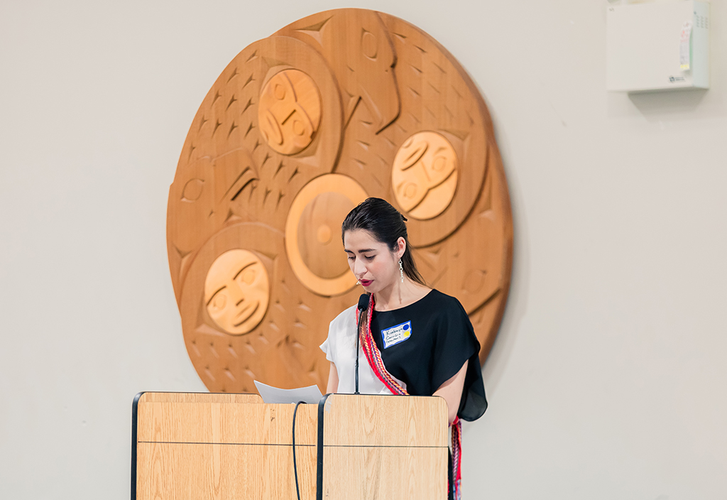 A young woman stands at a podium, giving a presentation. She is wearing a grey and white shirt; on top of this shirt, she wears a Métis sash.