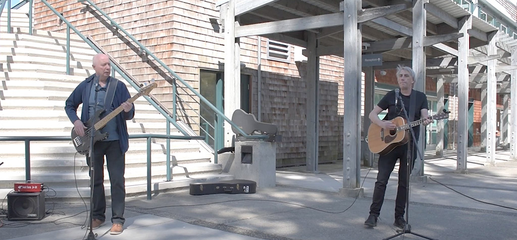 Two men stand outside of a community centre with their guitars plugged into amps in the middle of a song.