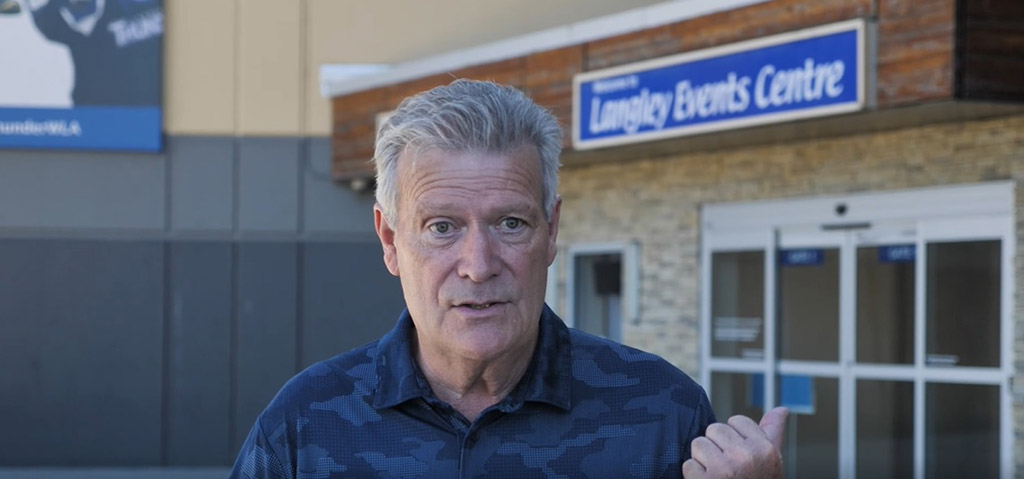 A white man in his mid-60s gestures to the Langley Events Centre behind him.