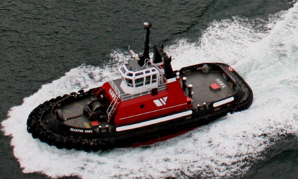 A black, red and white tugboat churns through the water, seen from above.