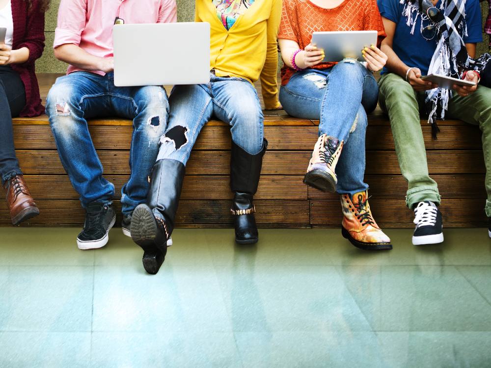 Picture features the legs of students sitting on a bench, one holds a laptop, the other an electronic tablet. 