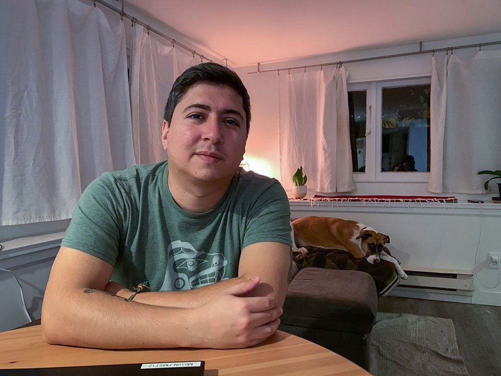 A man in his late-20s sits at the kitchen table of a humble home. It’s night and a lamp lights the scene. On the couch behind him is a dog.