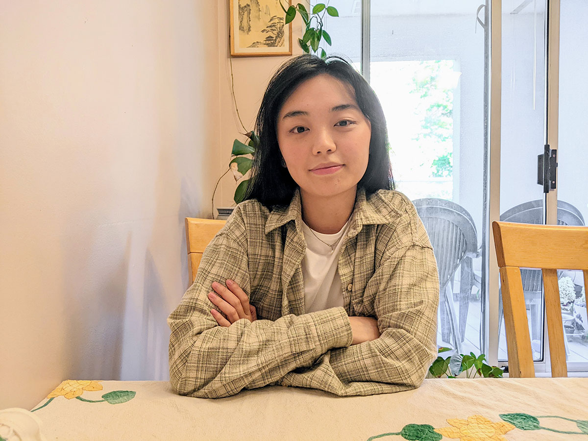 Wu sits at a kitchen table. There is a plant behind her growing towards the ceiling. On the wall is a small, framed work of Chinese watercolour art.