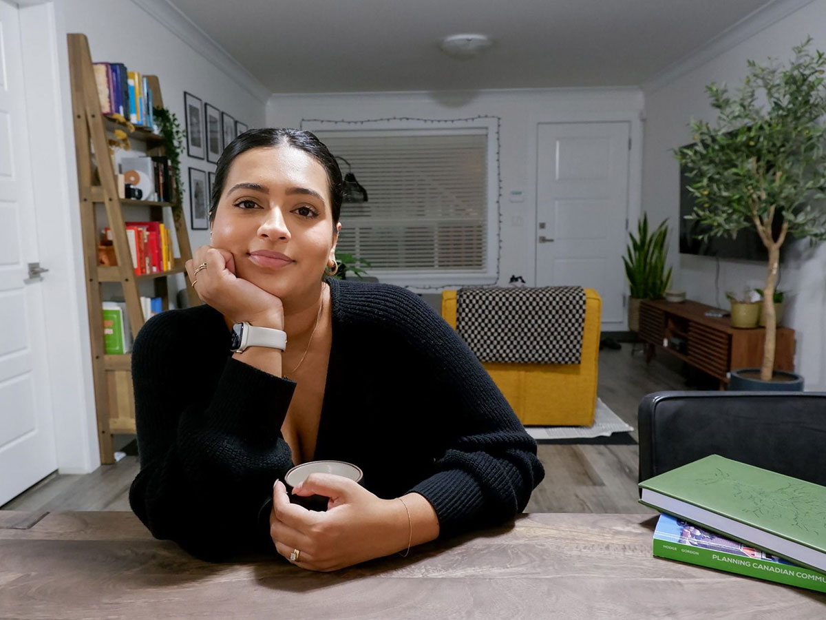 A woman who just turned 30 sits at her kitchen table. There is an urban planning textbook on it. We see that it is dark outside from the window. The living of her basement suite is behind her, with a bookshelf on one wall and a TV on the other. The home looks very new.