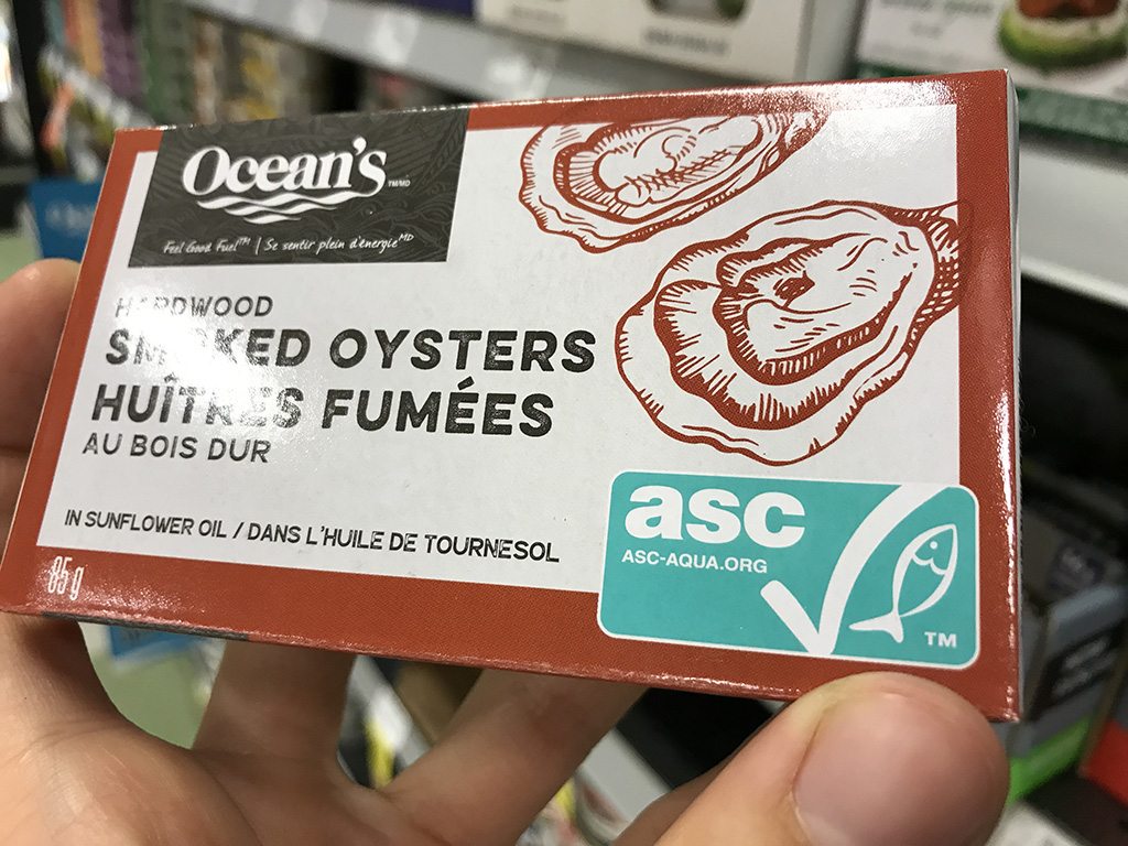 A hand holds a a package of oysters with a turquoise "asc" certification label in the bottom right corner.