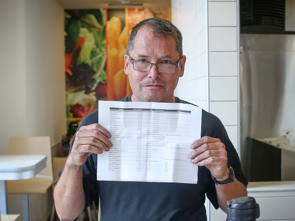 A middle-aged man in glasses and a dark blue shirt holds up a paper document.