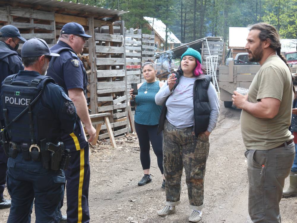 Three RCMP officers stand to the left. Four land defenders stand to the right. One is holding a cellphone, filming the interaction. 
