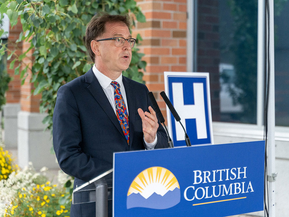 Adrian Dix, wearing glasses, a blue suit, white shirt and brightly patterned tie, stands behind a podi-um with a British Columbia sign. An "H" hospital symbol is behind him.