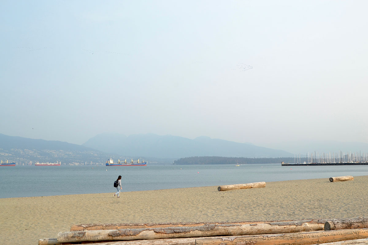 A view of Jericho Beach in Vancouver. There are logs resting on the sand in the foreground. A young woman is walking across the beach wearing a black backpack. The sky, hazy with wildfire smoke, is blue. Tankers are visible in the water.