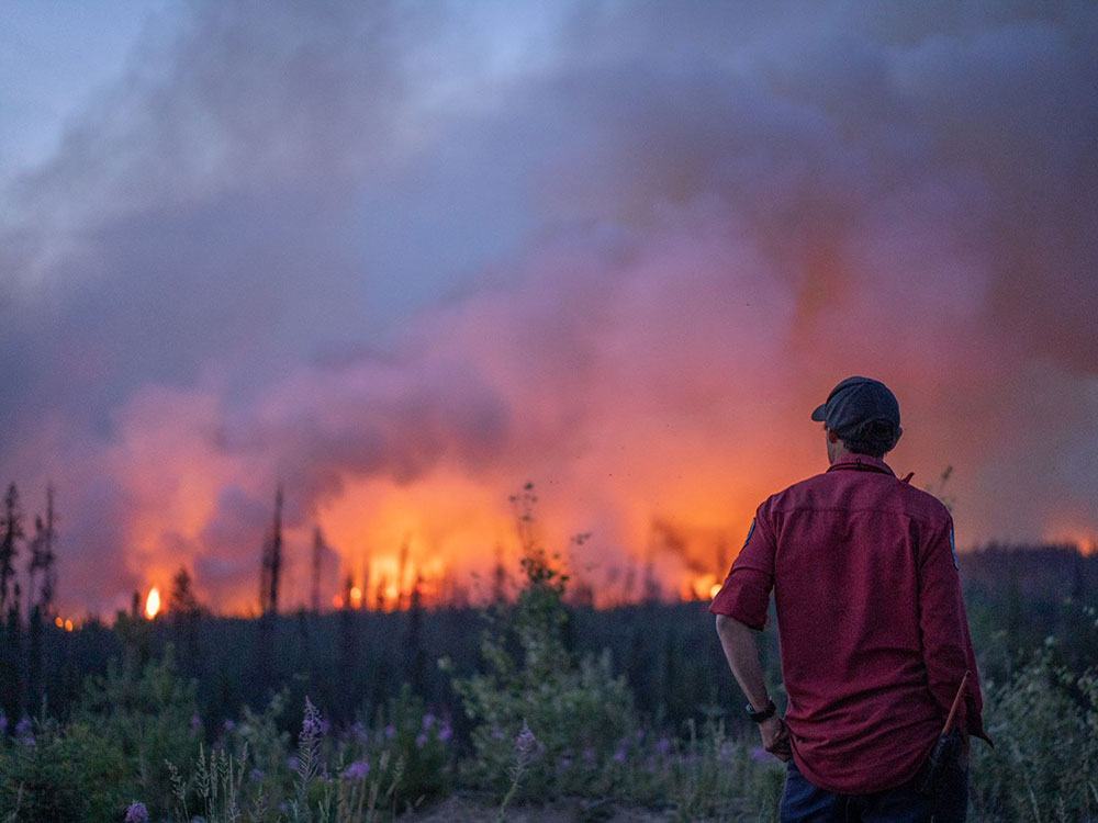 A man stands with his back to the camera. He looks towards a large wildfire in the background of the photo. 