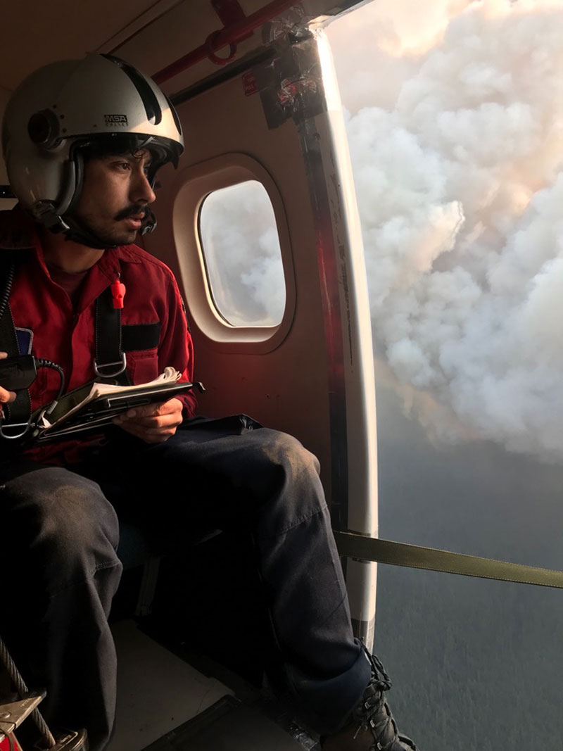 A man sits in a plane. The plane door is open. The man looks out at a wildfire below.