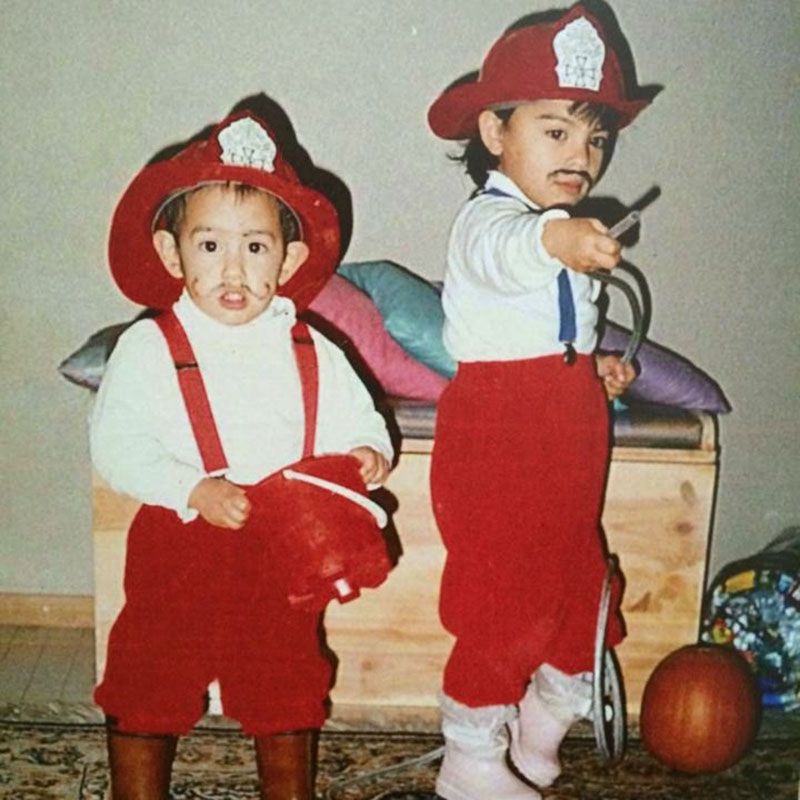 Two kids wear firefighter costumes. They look adorable and have moustaches drawn onto their faces.