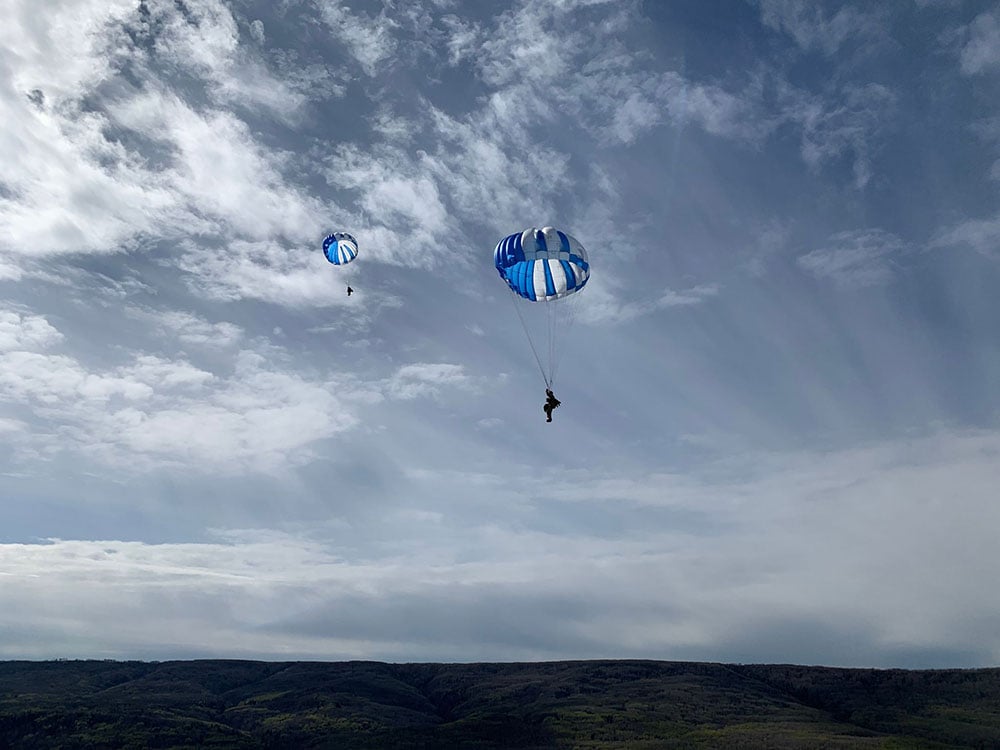 Two people float through the sky, harnessed into blue and white parachutes. Most of the photo is taken up by blue sky, but a hilly, treed horizon is visible in the bottom sixth of the photo.