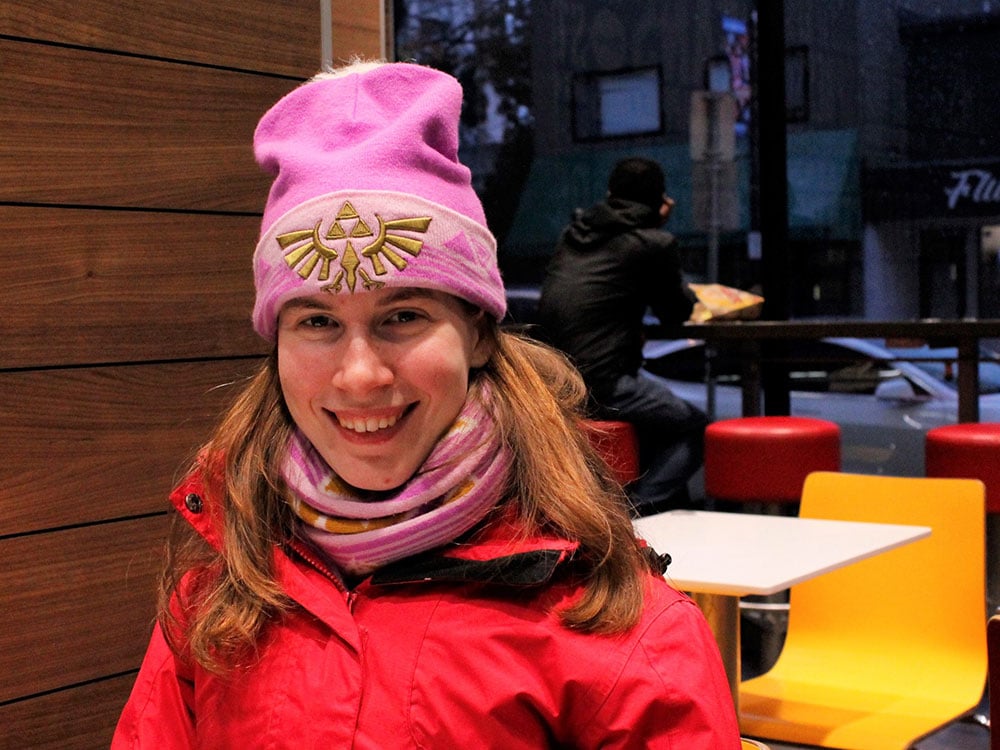 A woman wearing a fuchsia toque, scarf and red jacket sits in a room with vintage red and yellow seating, and wood panelling. She is smiling.