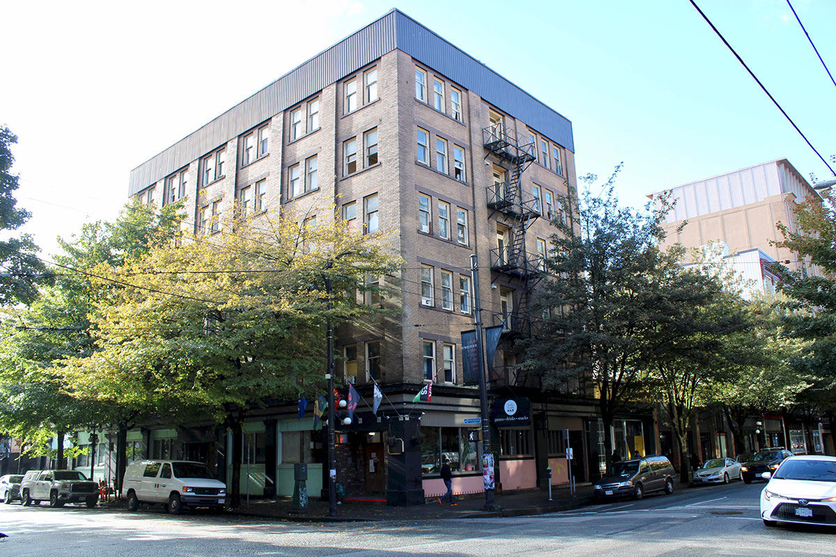 A six-storey building made of light-brown bricks with regularly spaced windows stands on a street corner, with trees in leaf in front.