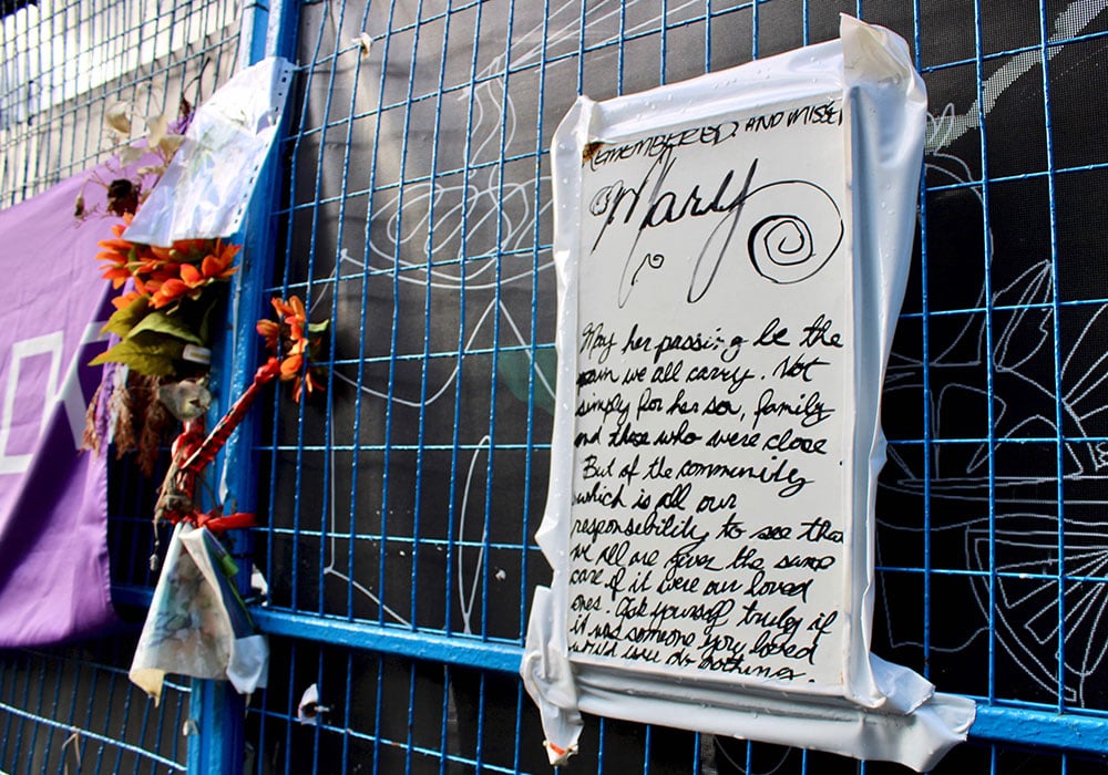 A closeup shot of a blue wire construction fence outside a black wall. On the right a purple banner and orange flowers are attached. On the left, a large white sign with black writing remembers Mary Ann Garlow, who died in the Winters Hotel fire. 