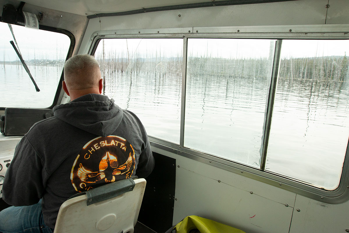 A man wearing a grey hooded sweatshirt that says “Cheslatta” on the back sits in the driver’s seat of a boat and looks out the window at the water. 