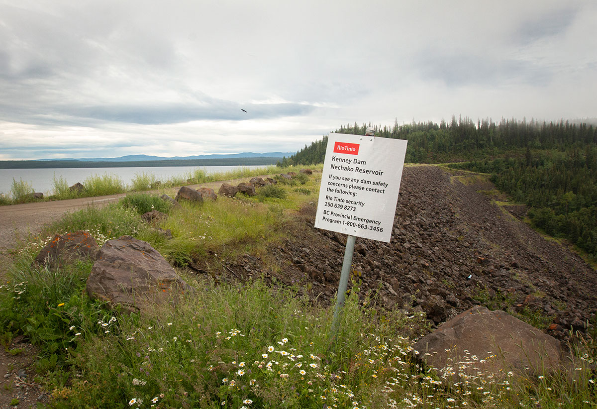 A reservoir stretches into the distance behind a grass-covered earth and rock dam with a dirt road running across its top. A sign asks people to report safety concerns.