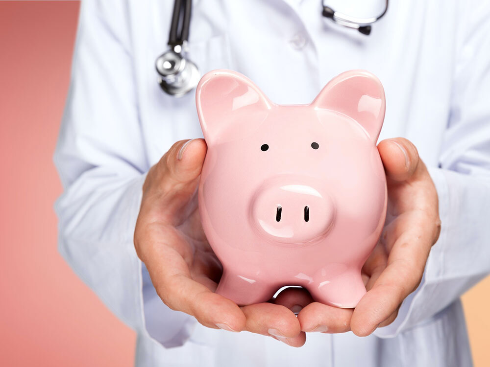 A person wearing a white coat and stethoscope holds a piggy bank out towards the camera.
